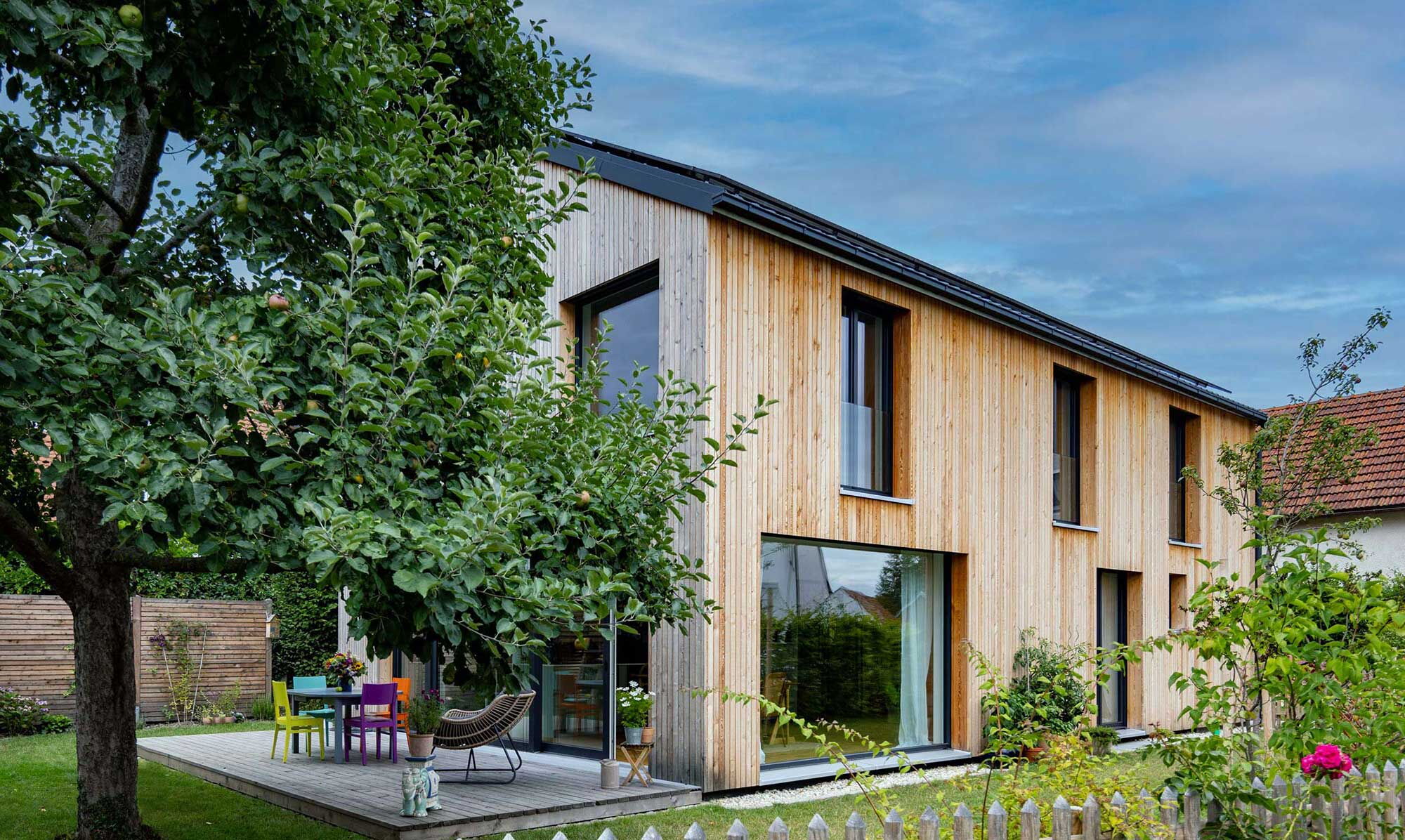 Ein modernes Holzhaus in Bayern steht inmitten eines grünen Gartens mit einem Apfelbaum und bunten Gartenmöbeln.
