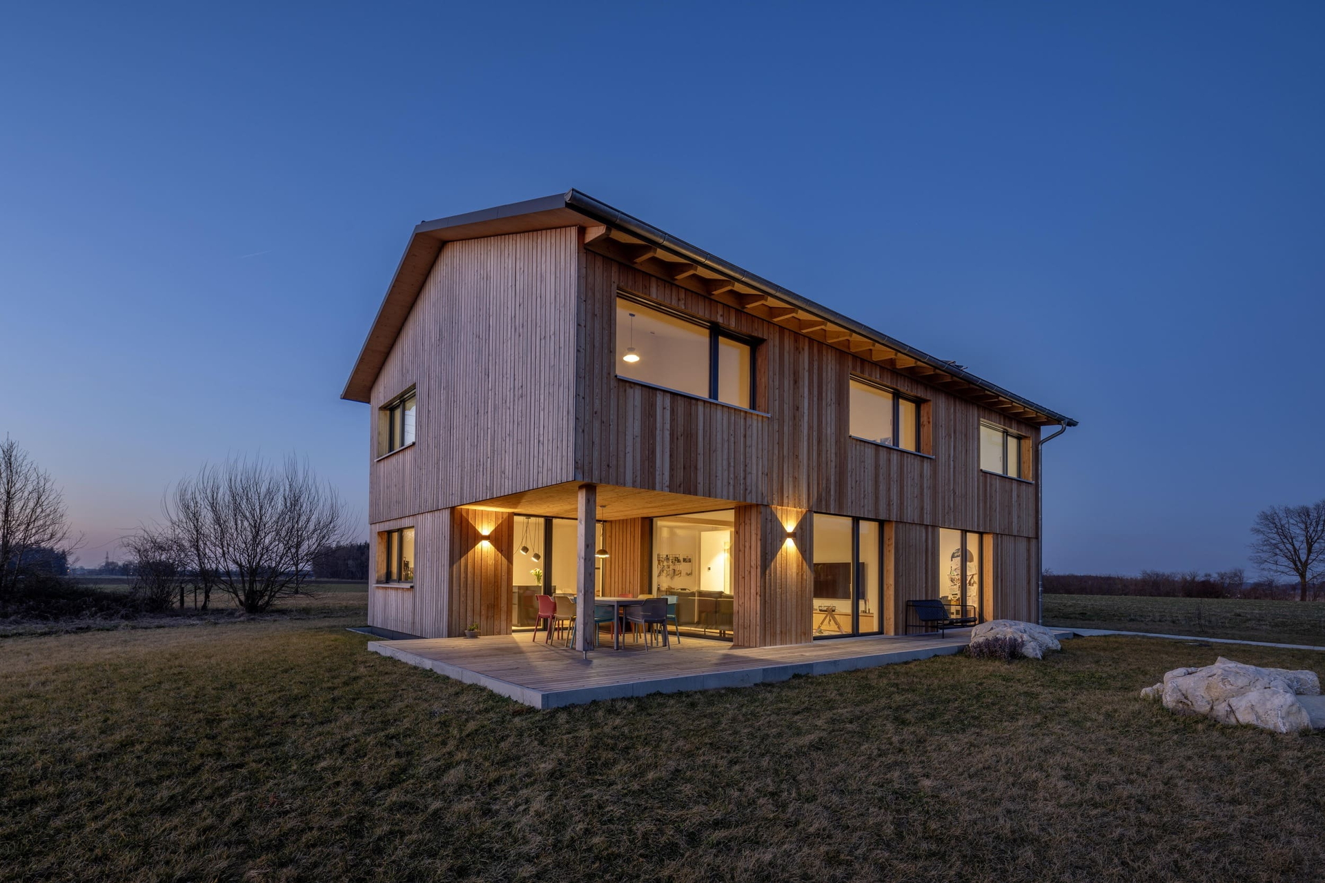 Ein modernes Holzhaus mit großen Fenstern leuchtet bei Dämmerung in einer offenen Landschaft mit klarer Architektur.