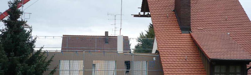 Ein Haus mit rotem Dach und Baumaßnahmen ist umgeben von einem Gerüst und einem großen Kran im Hintergrund.
