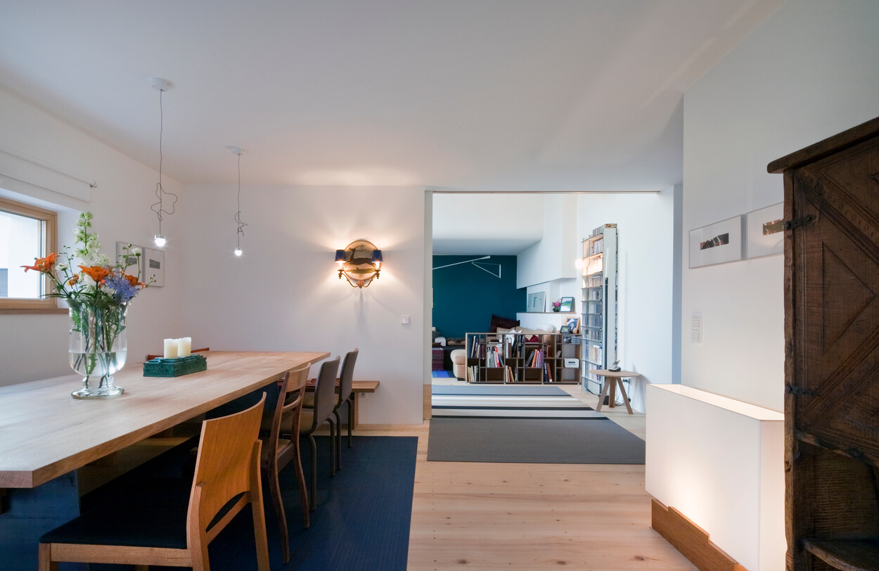 Ein modernes Esszimmer im Passivhaus Chieming mit Holztisch, blumiger Dekoration und einladendem Blick ins Wohnzimmer.