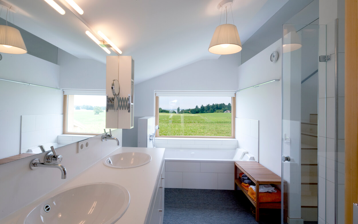 Modernes Badezimmer im Passivhaus Chieming mit zwei Waschbecken, großer Badewanne und Blick auf grüne Landschaft.