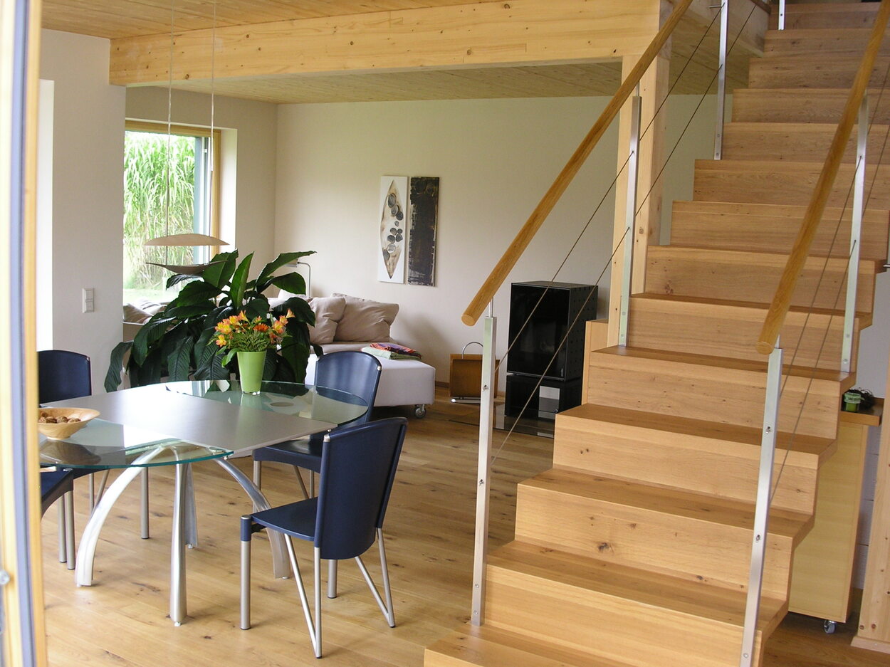 Ein modernes Interieur im Passivhaus-Stil mit Holztreppe, Glastisch und Pflanzen im hellen Wohnbereich.
