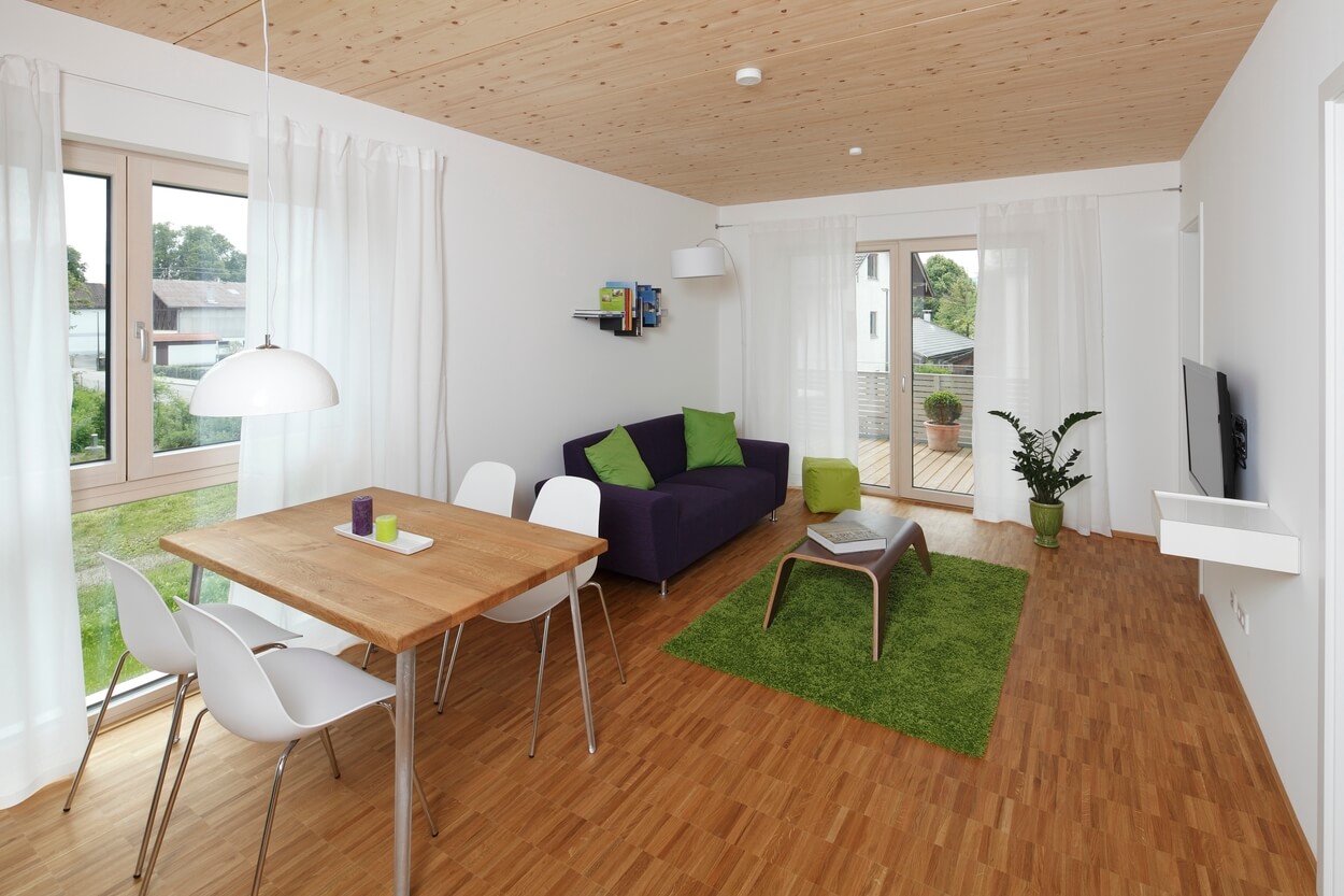 Ein modernes Wohnzimmer im Passivhaus mit Holzboden, grüner Deko und großen Fenstern, die viel Tageslicht hereinlassen.