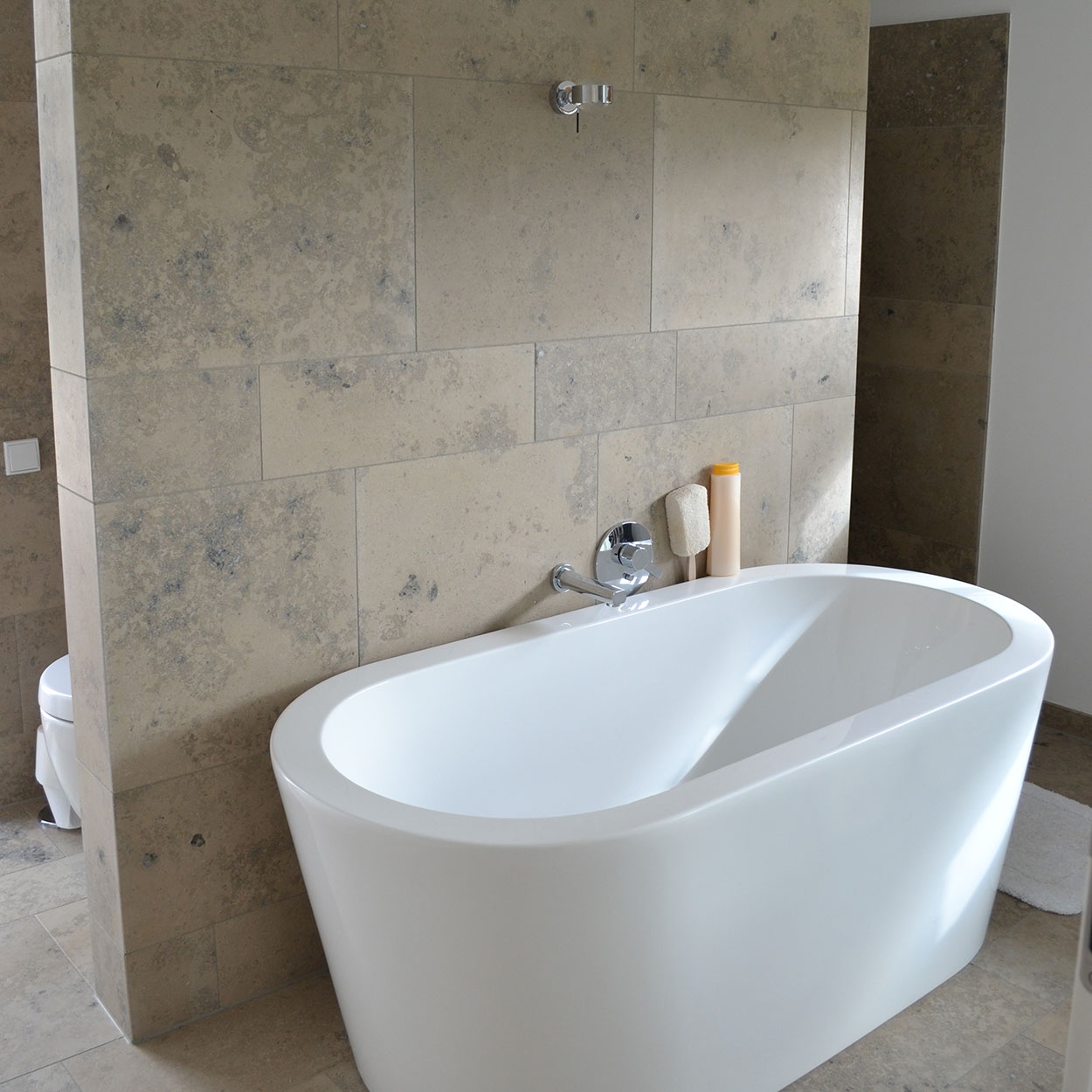 Ein modernes Badezimmer im Passivhaus-Stil zeigt eine freistehende weiße Badewanne vor beigen Wandfliesen.