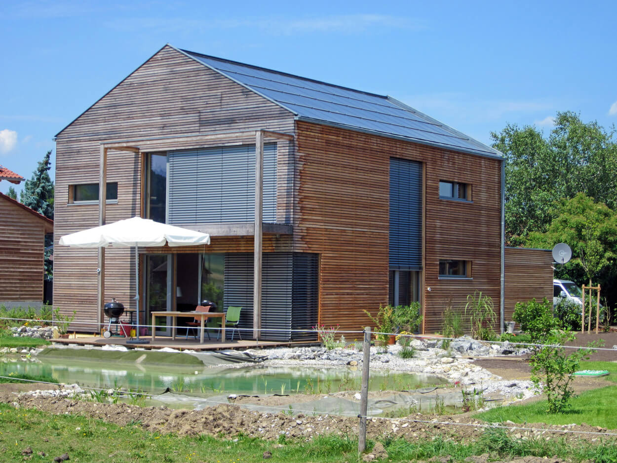 Ein modernes Passivhaus mit Holzverkleidung steht bei sonnigem Wetter neben einem kleinen Teich in Thalham.