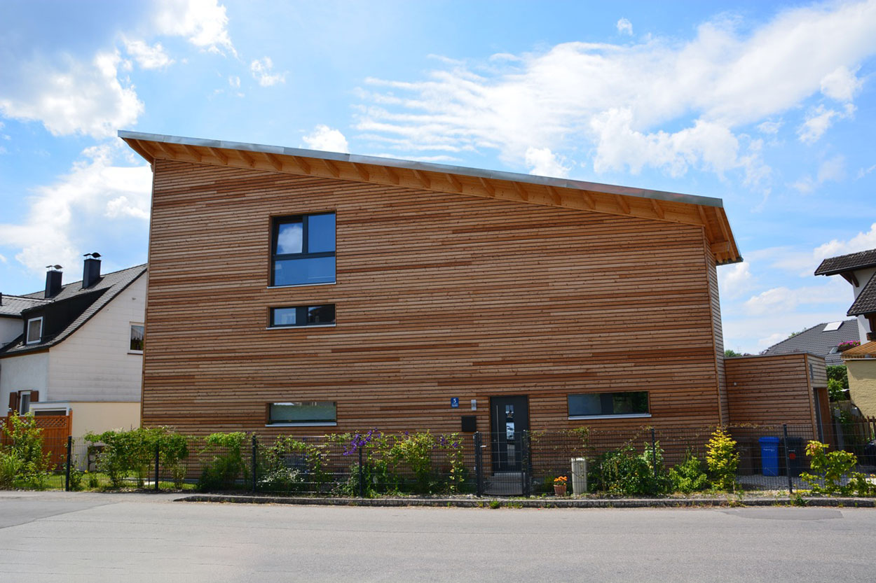 Ein modernes Passivhaus mit schrägem Dach aus Holz steht unter blauem Himmel in einem Wohngebiet in München.