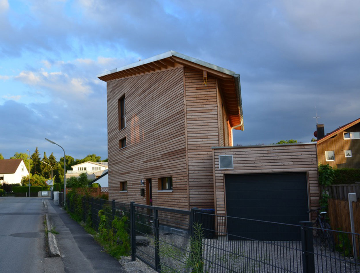 Ein modernes Passivhaus aus Holz steht in einer ruhigen Wohnstraße in München unter einem teils bewölkten Himmel.