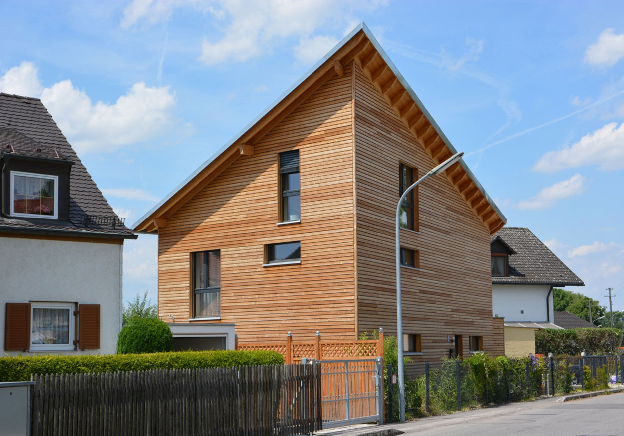 Ein modernes Passivhaus mit eleganter Holzverkleidung steht bei sonnigem Wetter in München neben traditionellen Häusern.
