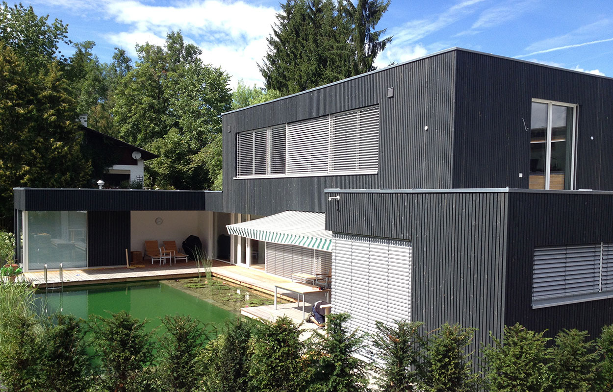 Ein modernes Passivhaus in Brannenburg mit dunkler Fassade steht in einem grünen Garten mit großem Naturteich.