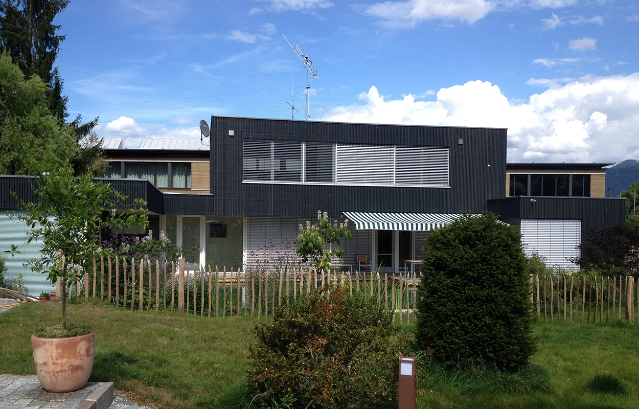 Ein modernes Passivhaus in Brannenburg steht vor einem klaren blauen Himmel, umgeben von grüner Natur und markanten Fenstern.