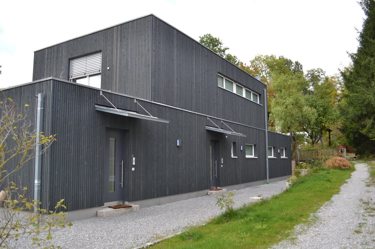 Ein modernes Passivhaus mit dunkler Holzfassade steht in Brannenburg, umgeben von üppiger grüner Natur.