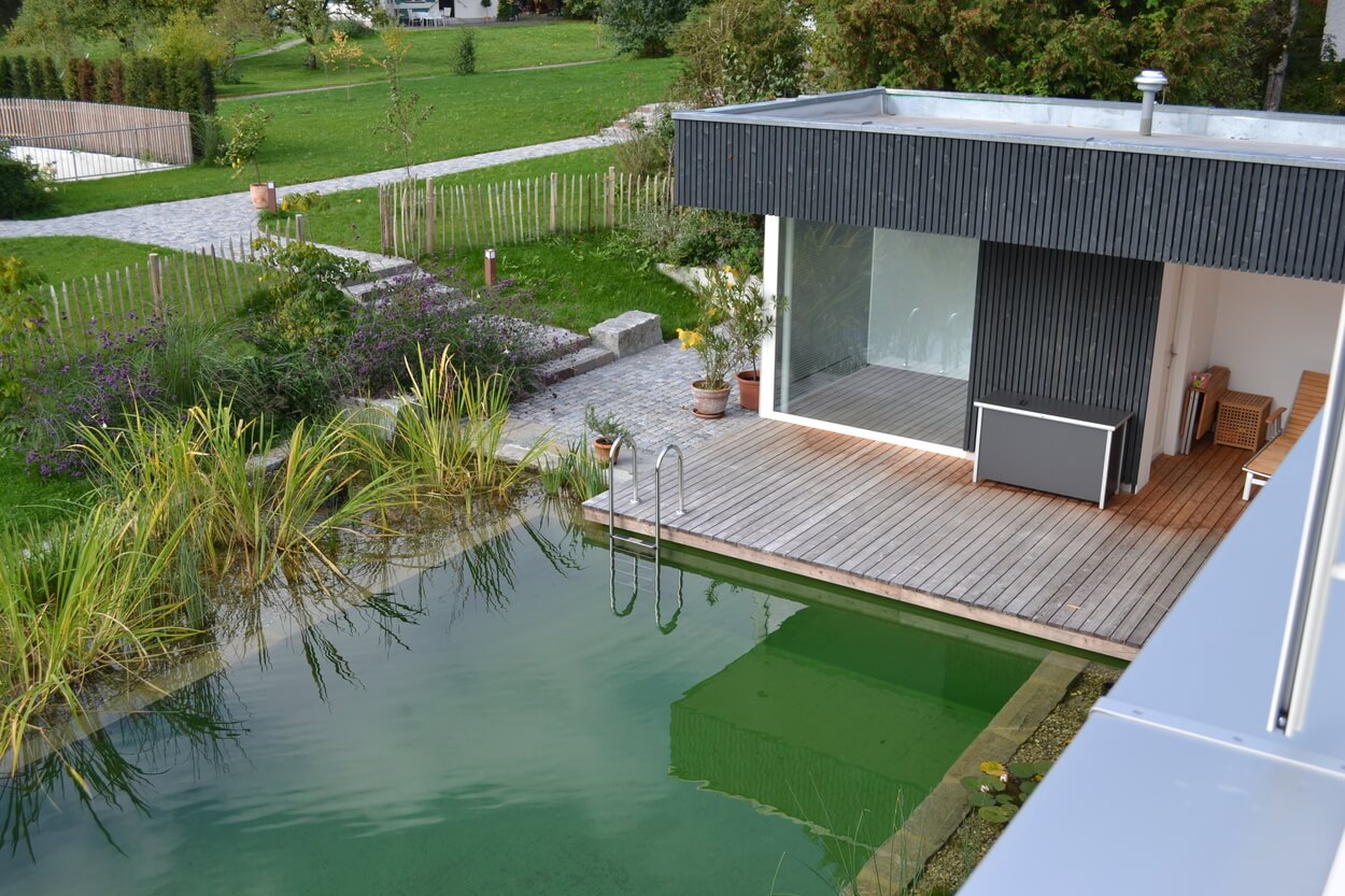 Ein modernes Passivhaus in Brannenburg mit Holzterrasse und natürlichem Schwimmteich liegt in grüner Umgebung.