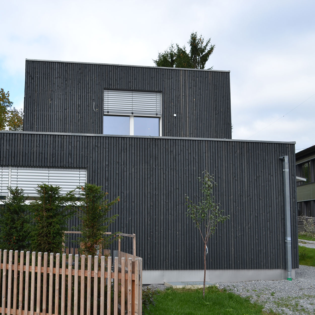 Das Passivhaus in Brannenburg besticht durch seine moderne, dunkle Holzfassade, einen kleinen Garten und einen Baum seitlich.