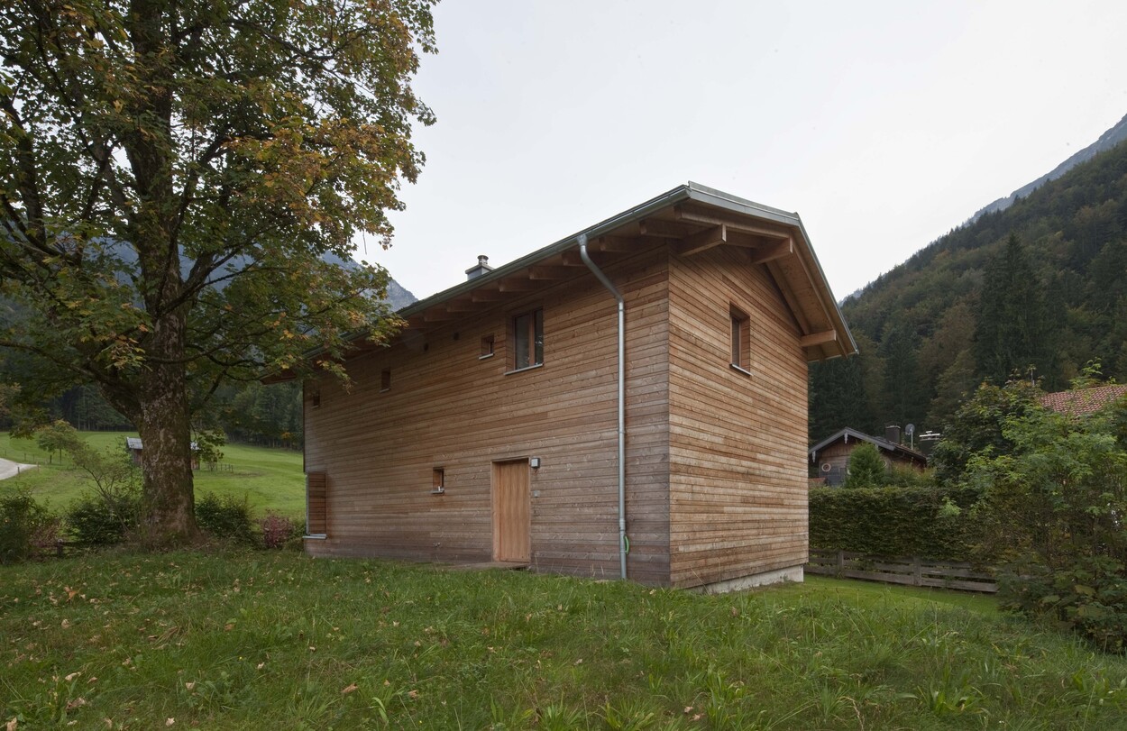 Ein schlichtes Holzhaus steht idyllisch inmitten grüner Wiesen und Bäume, umgeben von der malerischen Berglandschaft nahe Schliersee.