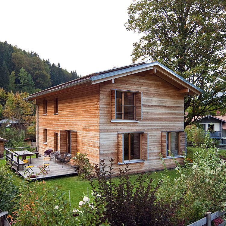 Ein modernes Holzhaus in Schliersee mit Terrasse steht idyllisch umgeben von grüner Natur und hohen Bäumen.