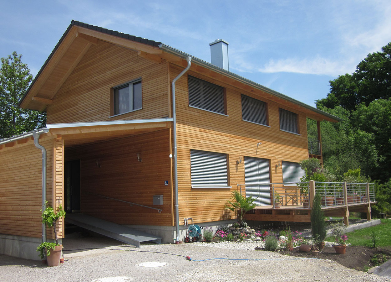 Ein modernes Holzhaus im Passivhaus-Stil in Glonn mit Terrasse und gepflegtem Garten strahlt bei sonnigem Wetter.