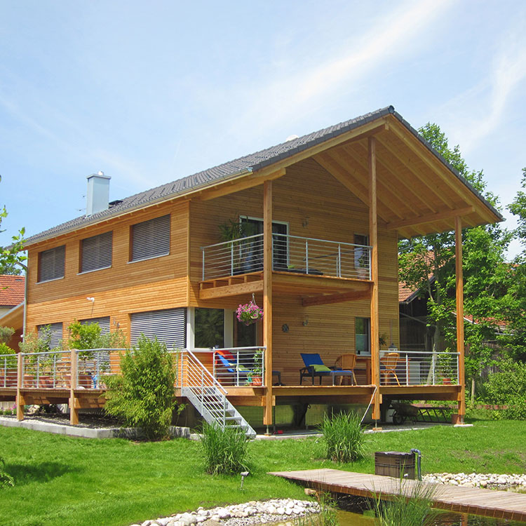 Ein modernes Holzhaus im Passivhaus-Stil steht in einem grünen Garten in Glonn, umgeben von Bäumen und einem Teich.