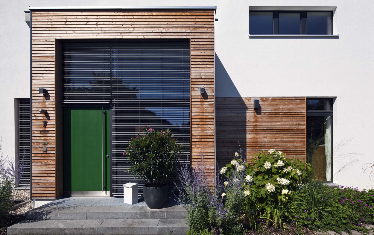 Ein modernes Passivhaus in Ismaning zeigt eine grüne Tür und Holzverkleidung, umgeben von üppiger Pflanzenpracht.