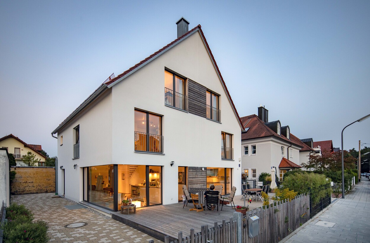 Ein modernes Doppelhaus in München mit beleuchtetem Innenraum und gemütlichem Außenbereich bei Abenddämmerung.