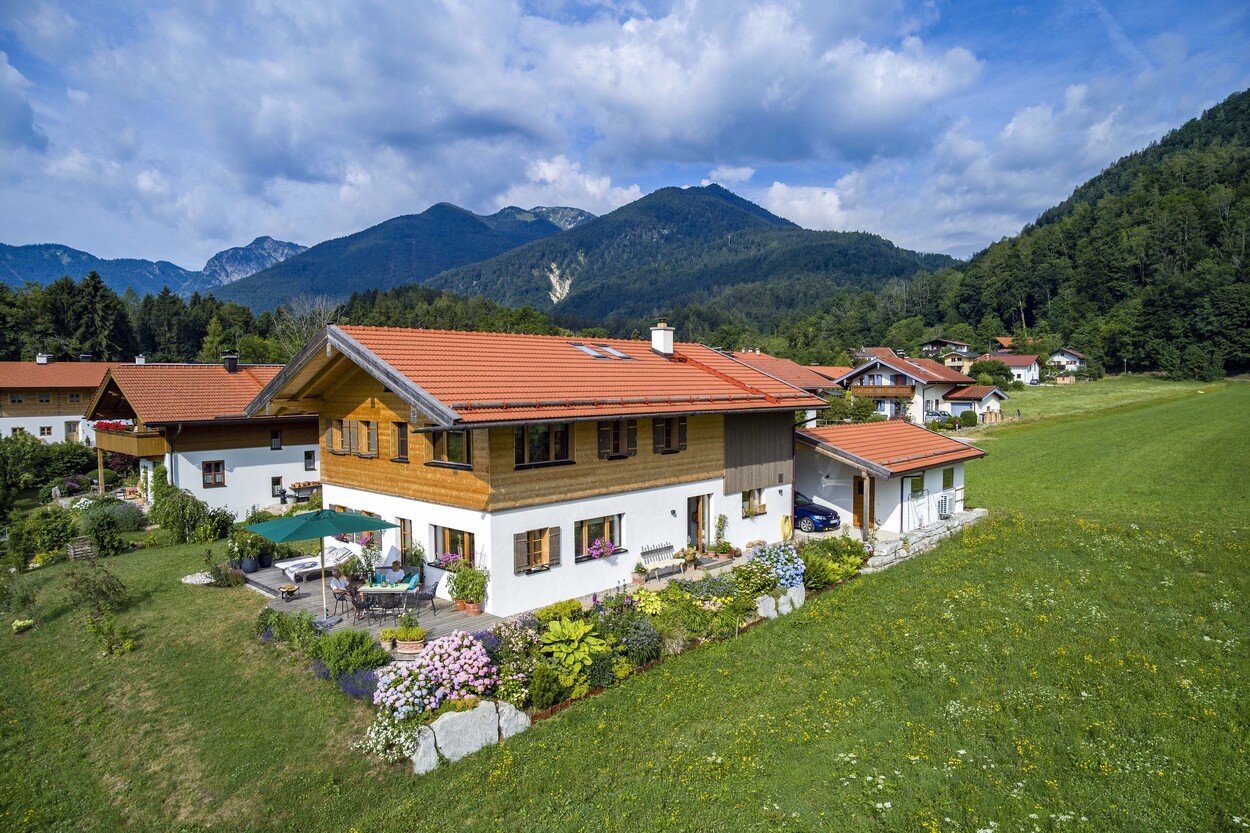 Ein traditionelles Holzhaus in Schleching steht malerisch vor einer Bergkulisse, umgeben von einem blühenden Garten.