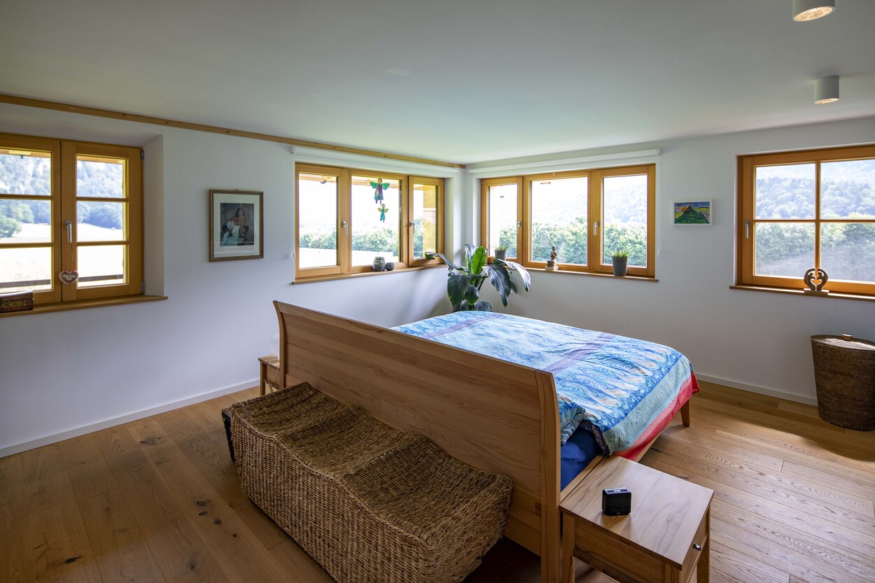 Ein gemütliches Schlafzimmer in einem Holzhaus in Schleching mit Holzmöbeln und großen Fenstern, die viel Licht hereinlassen.