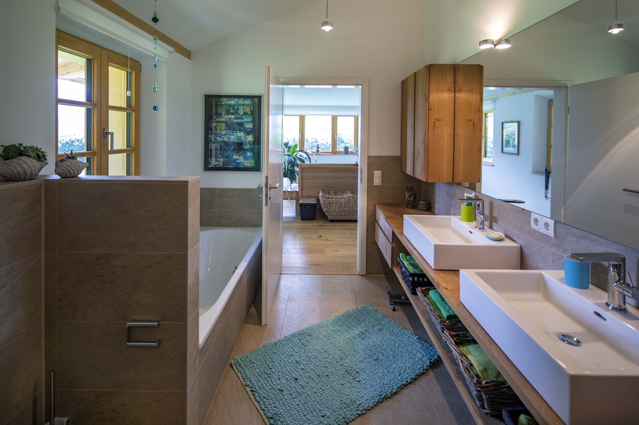 Ein modernes Badezimmer in einem Holzhaus in Schleching zeigt eine Badewanne, Doppelwaschbecken und elegante Holzmöbel.