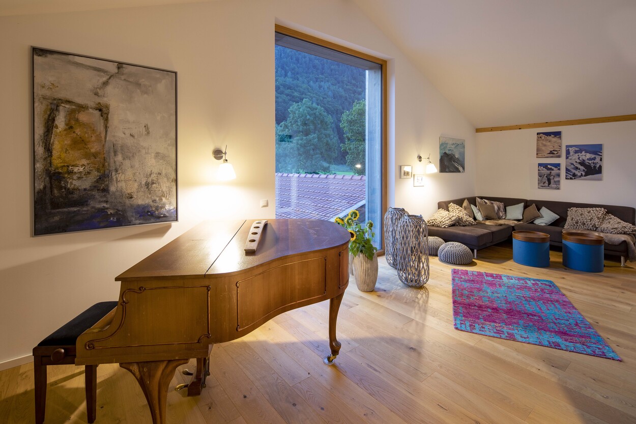 Ein stilvoller Raum in einem Holzhaus in Schleching mit Flügel, gemütlicher Sofaecke und großem Fenster mit Bergblick.