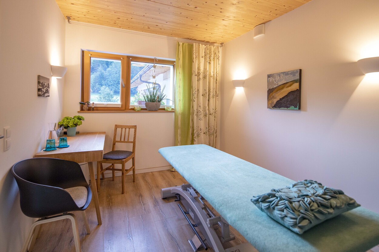 Ein heller Raum in einem Holzhaus in Schleching zeigt eine Massagebank, Pflanzen am Fenster und gemütliche Holzmöbel.