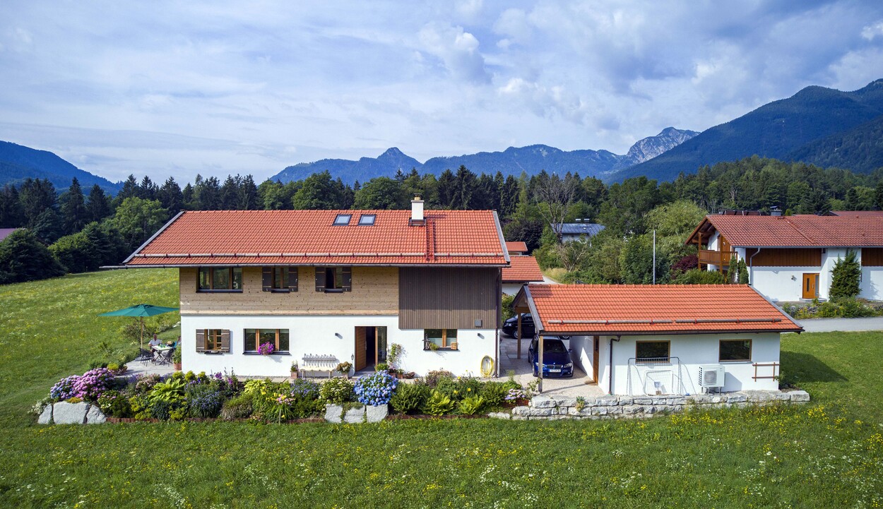 Ein charmantes Holzhaus in Schleching thront malerisch vor einer Bergkulisse, umgeben von grünen Wiesen und bunten Blumen.