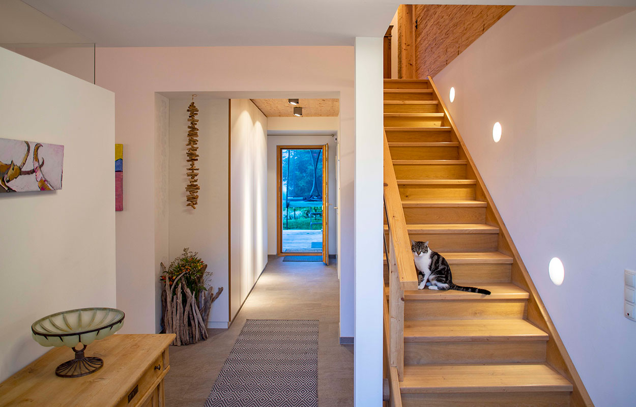Eine Katze sitzt auf einer Holztreppe im hellen Flur eines Hauses in Schleching, mit Blick auf den Garten.