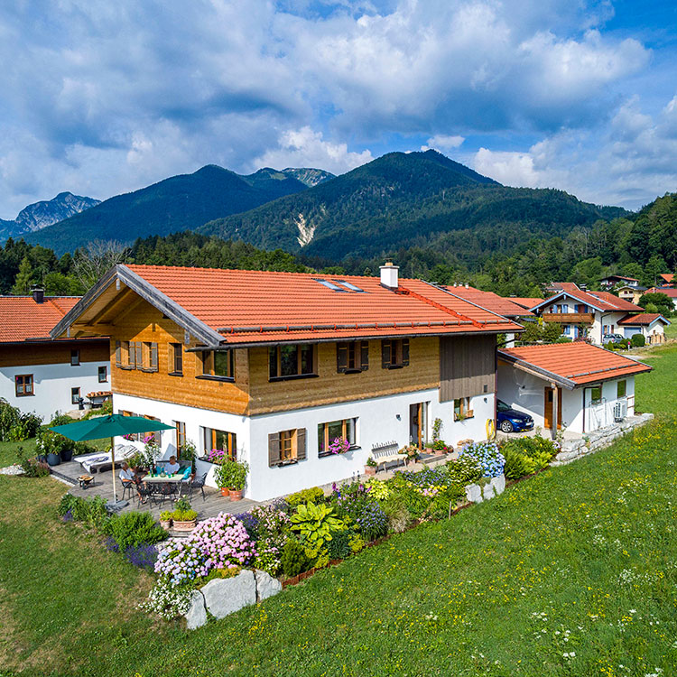 Ein charmantes Holzhaus in Schleching steht vor einer malerischen Bergkulisse, umgeben von blühenden Gärten und grünen Wiesen.