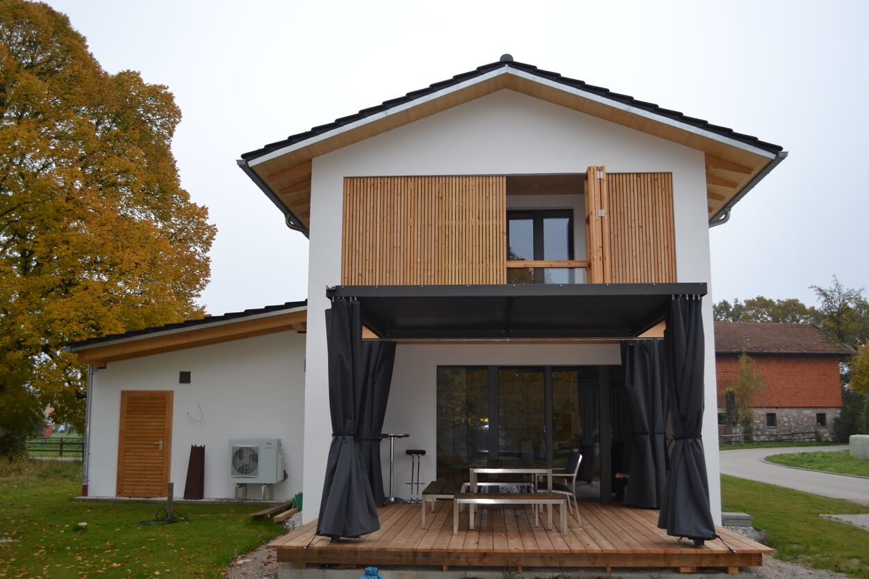 Ein modernes Passivhaus mit Holzfassade und Terrasse steht vor herbstlichen Bäumen in Übersee.