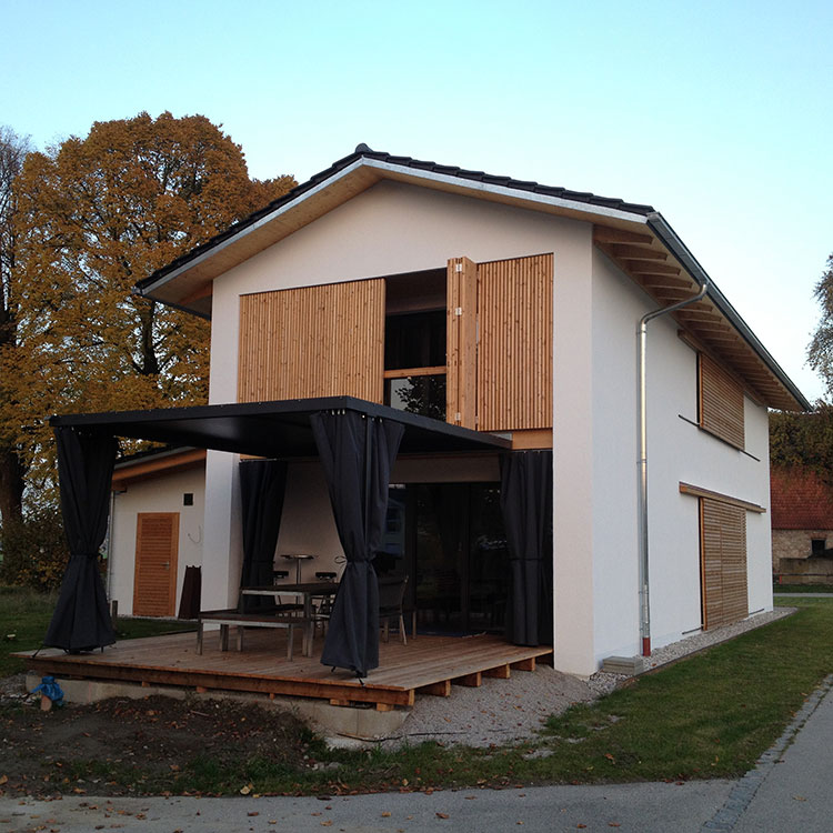 Ein modernes Passivhaus mit Holzverkleidung und überdachter Terrasse steht idyllisch in einer grünen Umgebung.