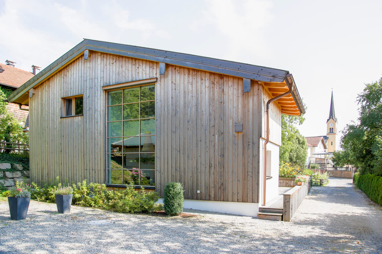Ein modernes Wohnhaus mit Passivhauskomponenten steht neben einem Kiesweg, während im Hintergrund eine Kirche sichtbar ist.