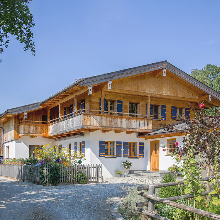 Ein traditionelles Wohnhaus mit Holzfassade und Balkon steht unter blauem Himmel, umgeben von einem blühenden Garten.