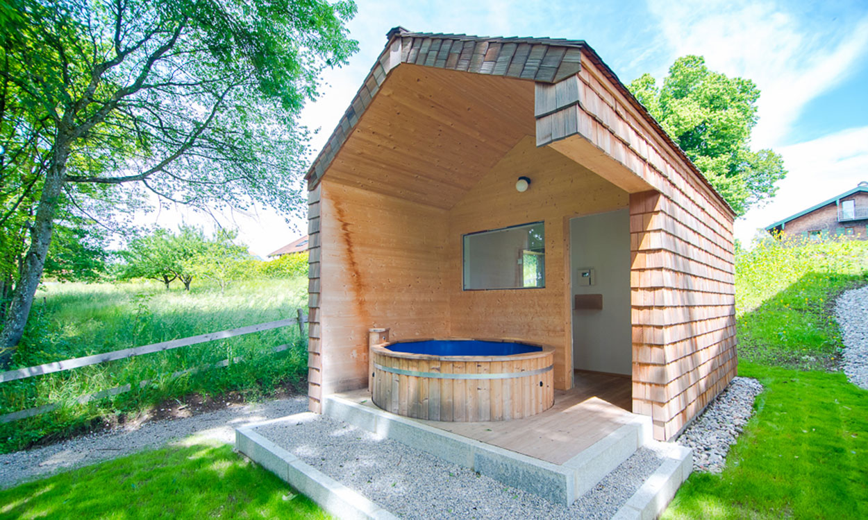 Ein modernes Passivhaus mit Holzverkleidung und Whirlpool steht idyllisch in einer grünen Landschaft unter blauem Himmel.