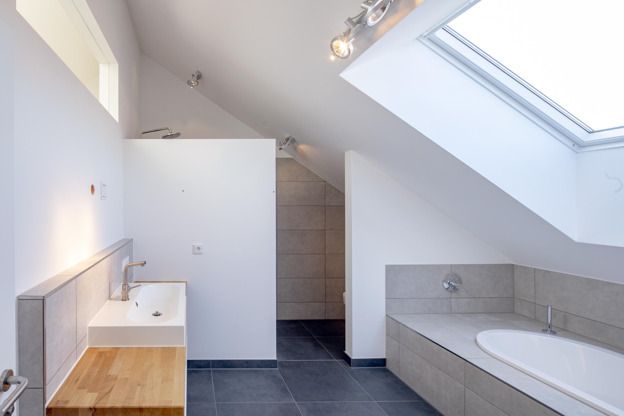 In einem modernen Badezimmer in Kirchseeon erhellt ein Dachfenster die Badewanne und den eleganten Waschtisch.