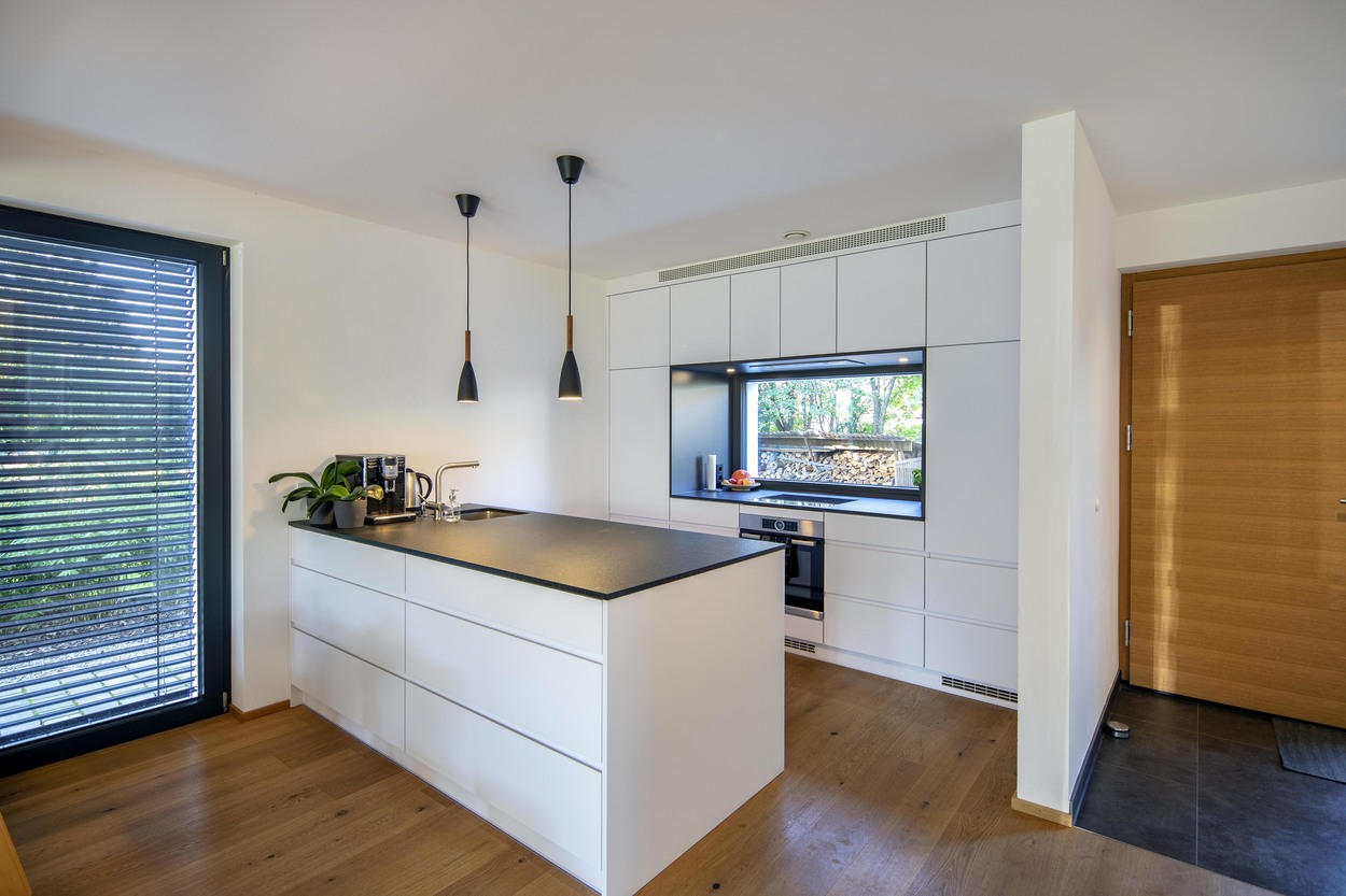 Eine moderne Küche in einem Mehrfamilienhaus in Kirchseeon mit weißer Insel, Holzfußboden und großem Fensterblick.