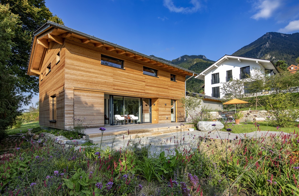 Ein modernes Holzhaus in Marquartstein, Chiemgau, steht von Garten und Bergen umgeben unter klarem, blauem Himmel.