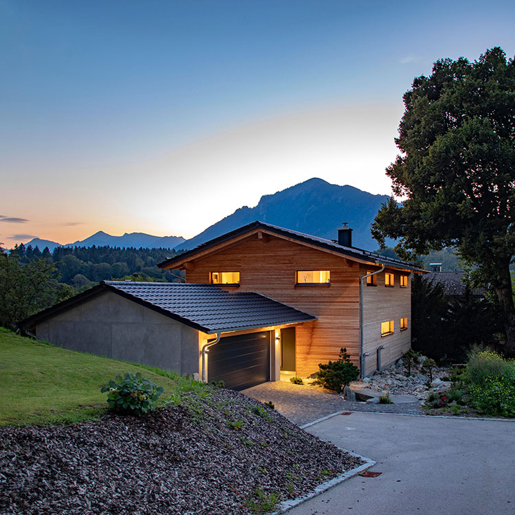 Ein beleuchtetes Holzhaus in Marquartstein steht malerisch vor der Alpenkulisse bei Sonnenuntergang.