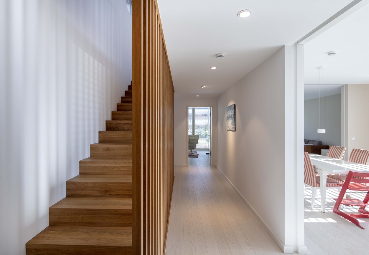 Ein moderner Flur in einem Passivhaus zeigt eine hölzerne Treppe und helle Räume mit roten Stühlen im Hintergrund.