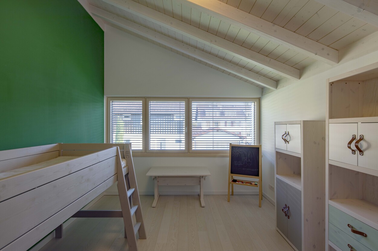 Ein helles Kinderzimmer in einem Passivhaus mit Hochbett, grünem Akzent und Tafel vor dem Fenster.