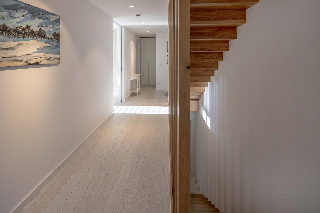 Das helle Treppenhaus eines Passivhauses in Andechs zeigt eine moderne Holztreppe und ein farbenfrohes Landschaftsgemälde.