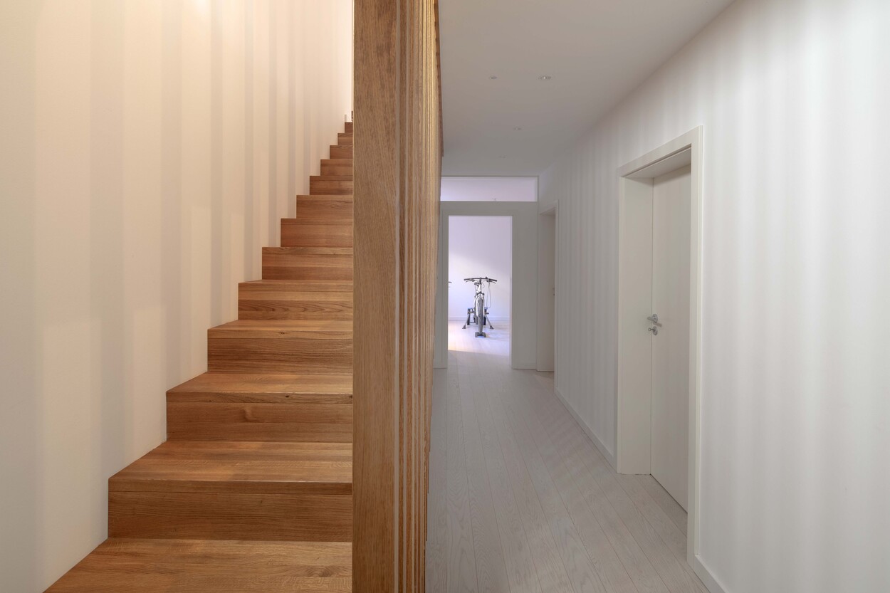Ein heller Flur im Passivhaus zeigt eine Holztreppe und ein Fitnessgerät im Untergeschoss von Andechs.
