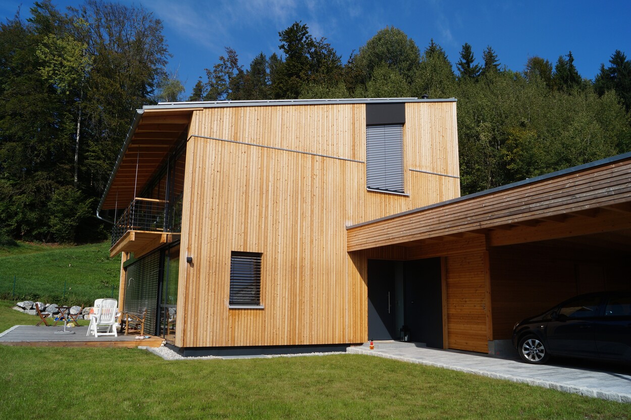 Ein modernes Einfamilienhaus aus Holz mit Terrasse und Carport in Feldkirchen, umgeben von grüner Natur.