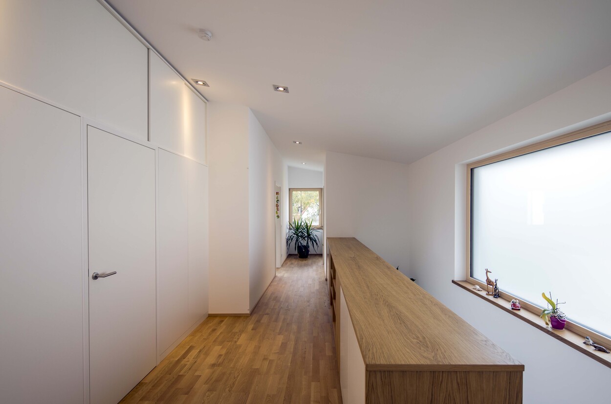 Ein heller Flur in einem Passivhaus in München mit Holzboden führt zu einem Fenster mit Pflanzen und minimalistischer Einrichtung.