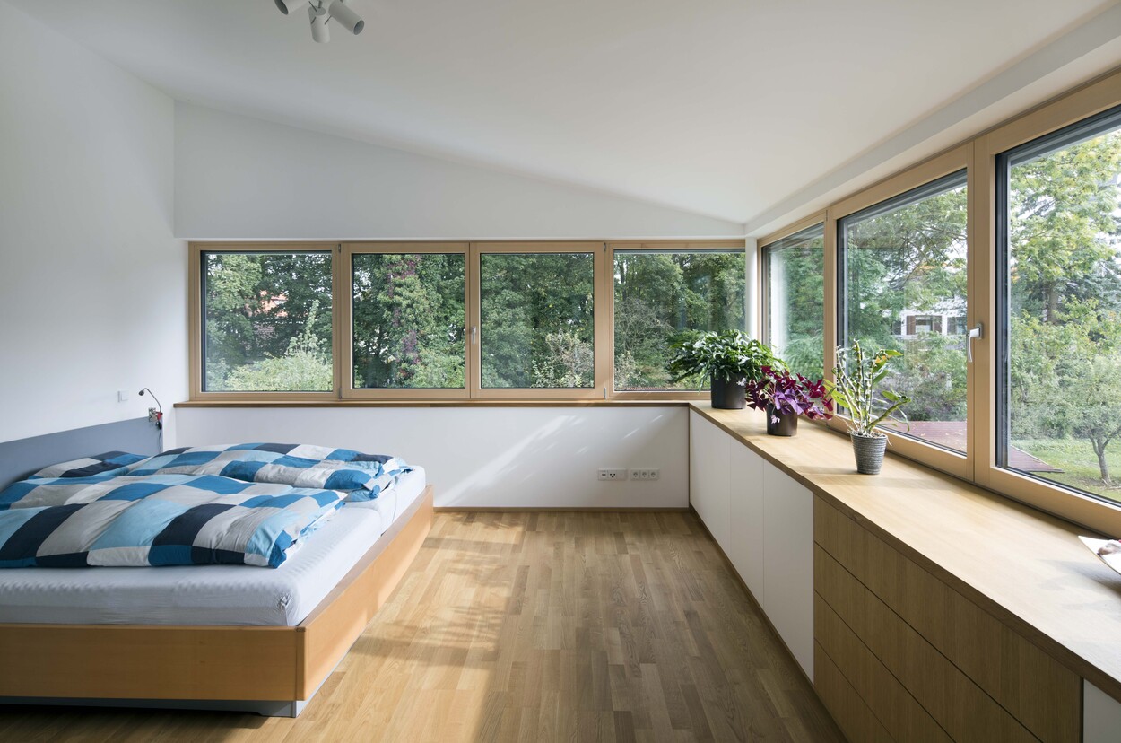 Ein helles Schlafzimmer in einem Passivhaus in München bietet durch große Fenster einen Ausblick auf grüne Bäume.