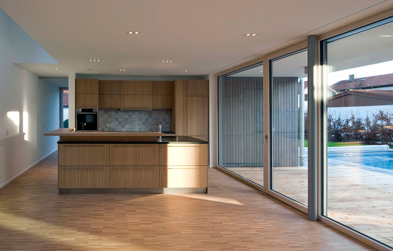 Eine moderne Küche im Passivhaus mit viel Tageslicht und Blick durch große Fenster auf den Pool in Bruckmühl.