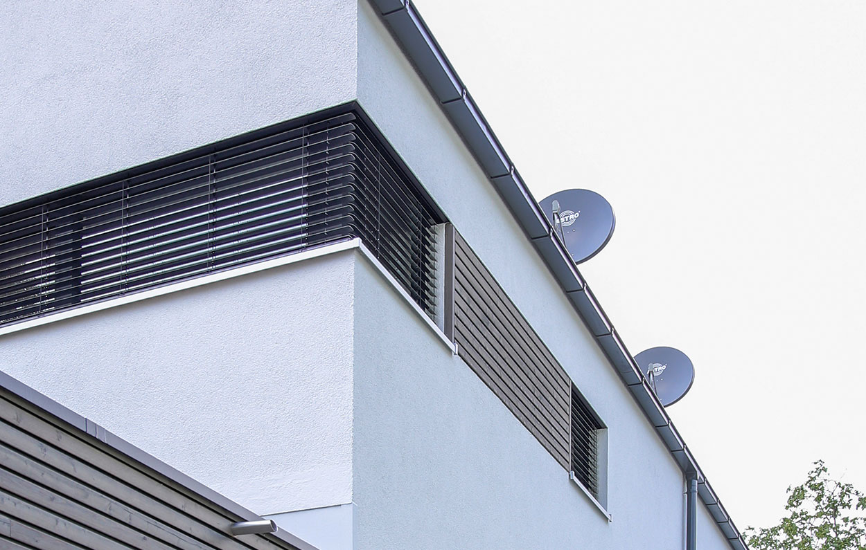 Ein modernes Passivhaus in München zeigt stilvolle Fenster mit Jalousien und drei Satellitenschüsseln auf dem Dach.