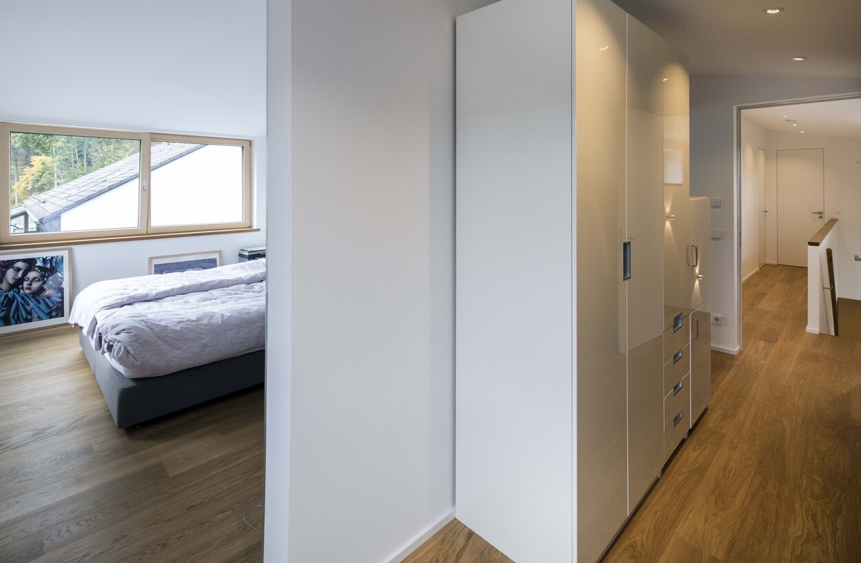 Ein heller Flur in einem energiesparenden Holzhaus in Putzbrunn führt zu einem luftigen Schlafzimmer mit großen Fenstern.
